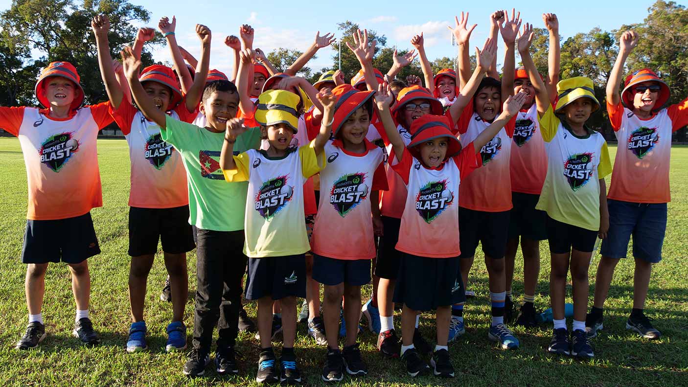 A group of primary school age children wearing a cricket uniform stand with their hands in the air